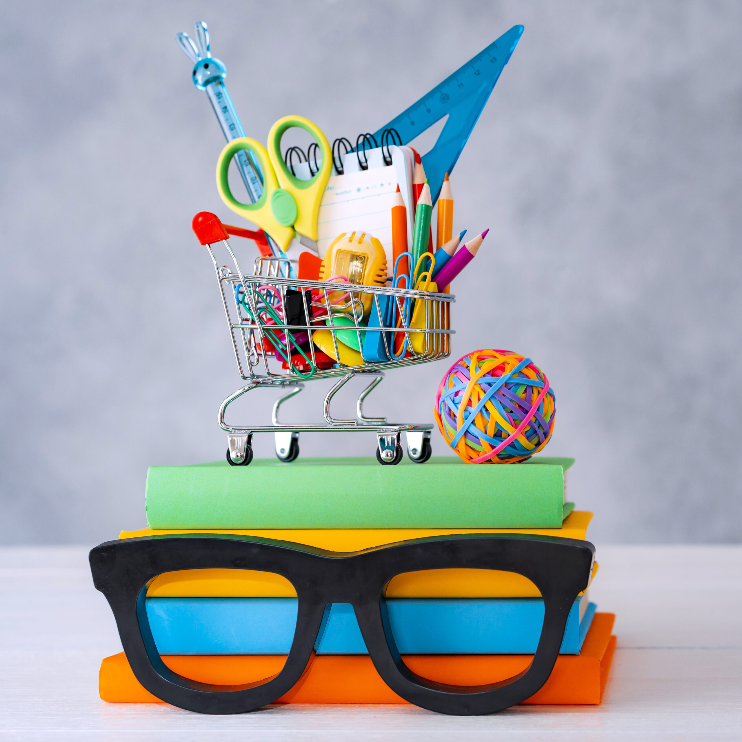 colorful school supplies shopping basket gray background with a copy text space. a stack of books with colorful covers frame glasses.